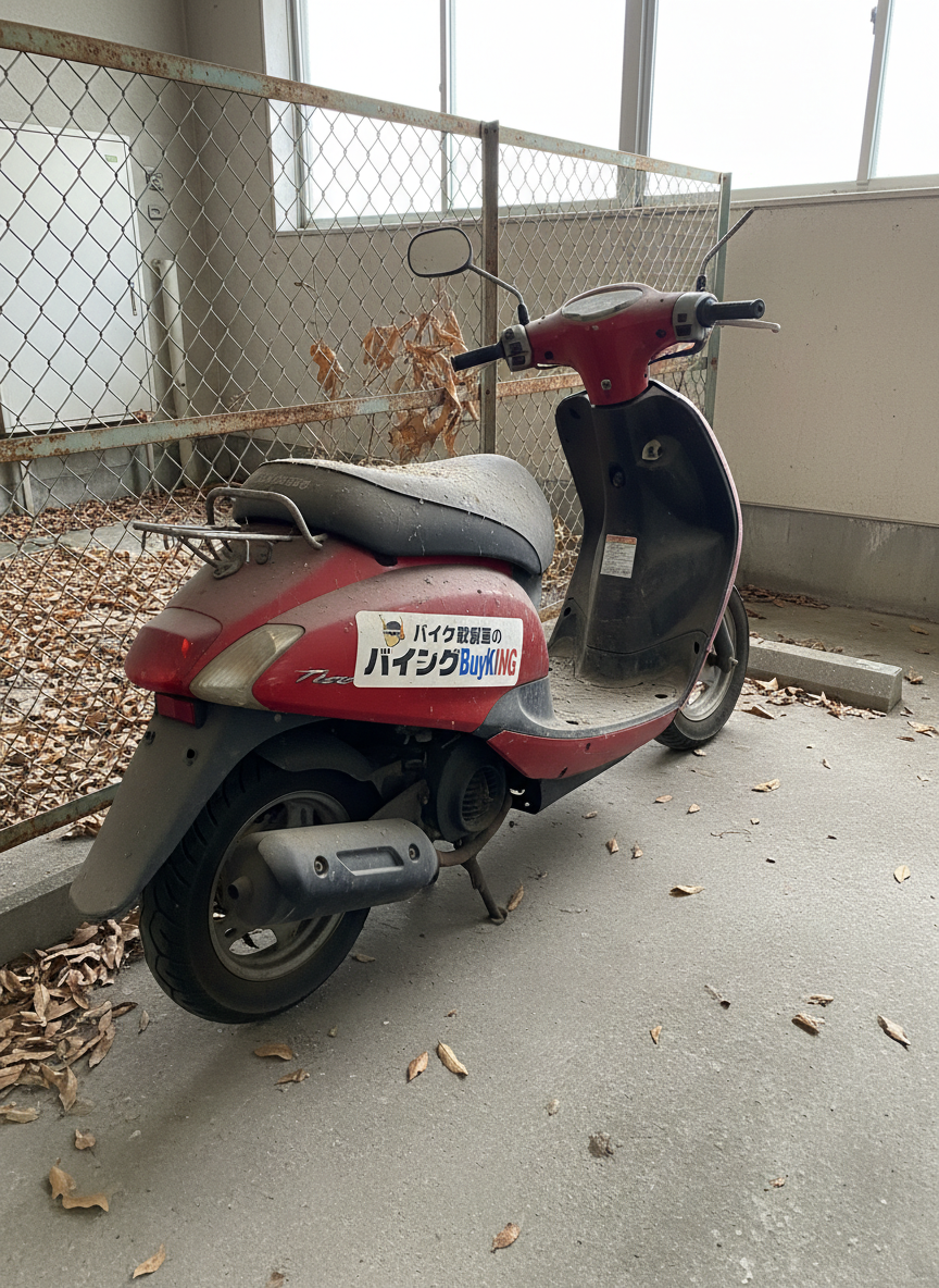 A neglected, dust-covered scooter with flat tires and faded red paint sitting in a cramped apartment parking space, surrounded by scattered dry leaves and a slightly rusted metal fence. A subtle branded sticker of "バイク買取のバイキングBuyKING" is affixed to the scooter’s side panel, suggesting it is awaiting pickup. Overcast daylight from a cloudy sky provides soft, even illumination with muted shadows, emphasizing the texture of dust, rust, and worn rubber without looking harsh. Photographic realism, captured at eye level with moderate depth of field so both the scooter and immediate environment are clearly visible. The atmosphere is calm yet slightly melancholic, conveying long-term neglect but also the reassuring possibility of proper disposal and purchase despite the condition.